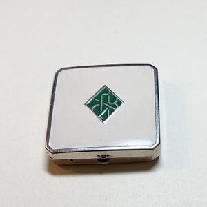 May include: A rectangular, silver-toned compact with a white enamel top. A green, diamond-shaped emblem with an intertwined design is centered on the lid. The compact has a hinged closure and a polished finish.