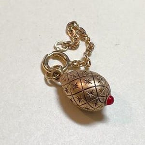 May include: A gold-colored charm with a textured, egg-shaped design and a small red accent. The charm is attached to a gold chain and a circular clasp. The egg-shaped portion has a grid pattern with star-shaped details.