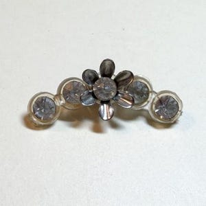 May include: A hair clip featuring a central metallic flower with six petals and a clear circular rhinestone at its core. The design is flanked by two clear circular rhinestones on each side, all set within a transparent plastic frame.