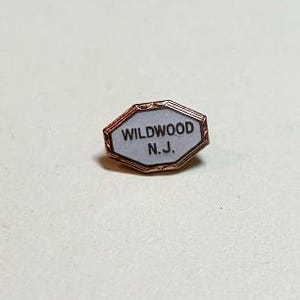 May include: A small, copper-toned pin with an octagonal shape. The pin features the text "WILDWOOD N.J." in black on a white background, framed by a thin copper border. The pin is likely a souvenir or collectible item.