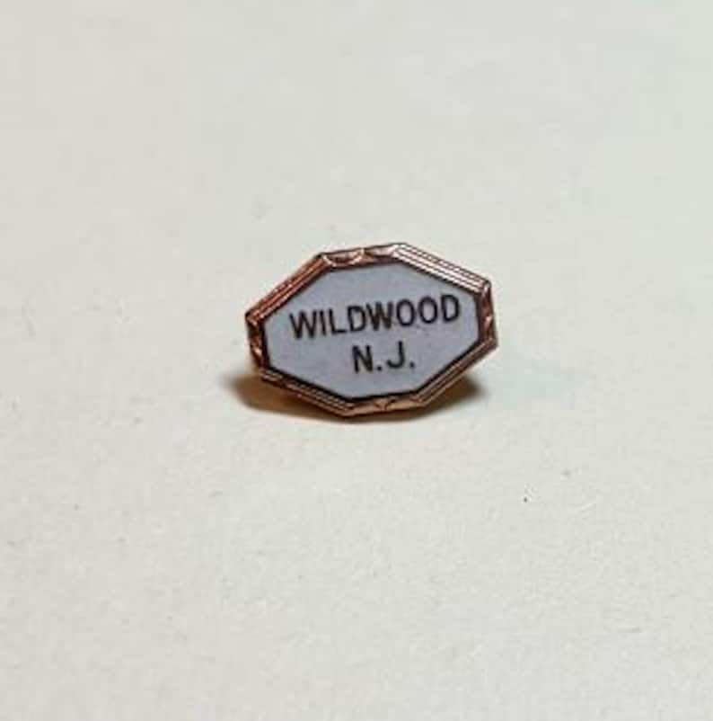 May include: A small, copper-toned pin with an octagonal shape. The pin features the text "WILDWOOD N.J." in black on a white background, framed by a thin copper border. The pin is likely a souvenir or collectible item.