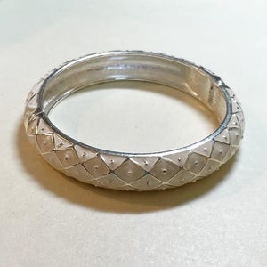 May include: A silver-toned hinged bangle bracelet with a geometric, diamond-patterned design. The bracelet features a light pink enamel inlay and small, raised dots at each intersection of the pattern. The bracelet is a fashion accessory.
