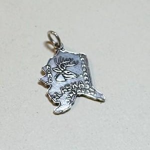 May include: A silver-toned metal charm shaped like the state of Alaska. The charm features an embossed graphic of a deer head, the word ALASKA, and the text NOME and FAIRBANKS printed along the edges. A small circular jump ring is attached to the top.