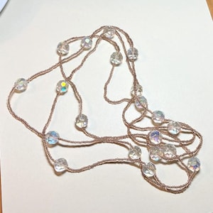 May include: A long, layered necklace featuring iridescent, clear crystal beads strung on a delicate, brown cord. The beads are interspersed along the cord, creating a flowing, elegant design. The necklace is displayed on a neutral background.