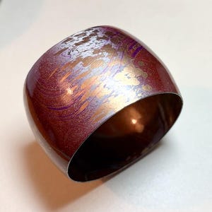 May include: A wide, curved cuff bracelet with a metallic finish. The bracelet is primarily burgundy with splashes of gold, silver, and purple. The interior of the bracelet is a warm copper color. The bracelet has a smooth, polished surface.