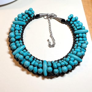May include: A statement necklace featuring a dense arrangement of turquoise-colored beads in various round and cylindrical shapes, interspersed with smaller black beads. The piece includes a silver-toned metal clasp and an adjustable chain extender.