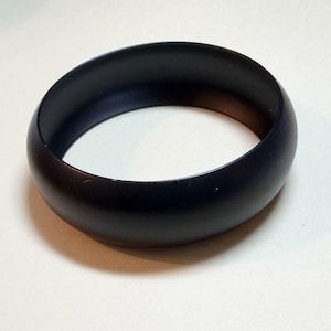 May include: A solid black bangle bracelet. The bracelet is a thick, rounded band with a smooth, matte finish. The dark color and simple design make it a versatile accessory.