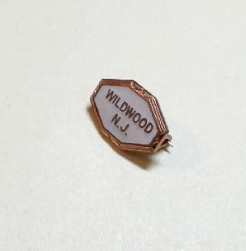May include: A vintage Wildwood N.J. pin with a copper-colored frame and a white background. The pin is in the shape of an irregular octagon. The text "WILDWOOD N.J." is printed in black on the white background.