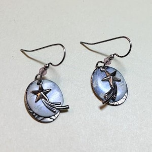 May include: A pair of silver-toned earrings. Each earring features a light blue oval pendant with a star and a shooting star design. The earrings have a hook closure and a small pink bead.