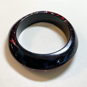 May include: A dark, glossy bangle bracelet with a black interior and a swirling, speckled exterior in shades of red, blue, and black. The bracelet has a smooth, rounded shape.
