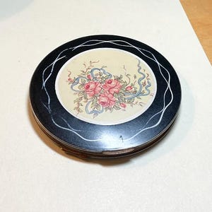 May include: A black compact with a white floral design on the lid. The floral design features pink roses and blue ribbons.