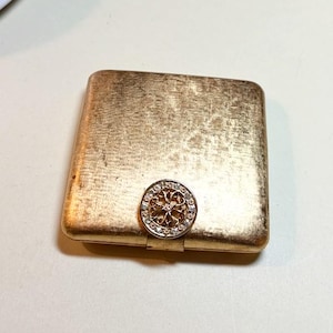 May include: A rectangular, gold-colored compact with a textured surface. The compact features a decorative, round clasp with a floral design and small, clear gemstones. The clasp is silver-colored.