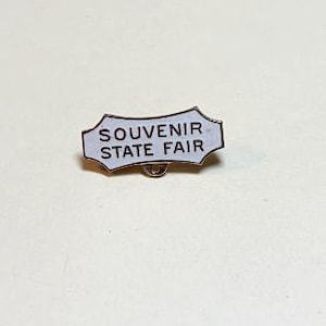 May include: A small, white enamel pin with the text "SOUVENIR STATE FAIR" in black lettering. The pin has a silver-colored border and a pin back. The pin is likely a souvenir item.