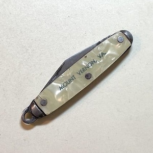 May include: A vintage pocket knife with a pearlescent handle. The handle is cream-colored and features the text "MOUNT VERNON, VA" in dark green. The knife has a silver-colored blade and metal accents, including a loop for attaching a lanyard.