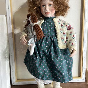 May include: A porcelain doll with auburn curly hair, wearing a green floral dress, a cream-colored cardigan with floral accents, and beige shoes. The doll holds a smaller doll. The Hamilton Collection logo is visible.