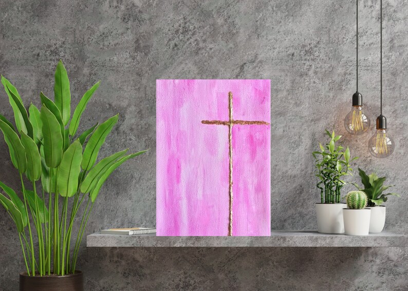 May include: A pink canvas painting with a gold cross in the center. The cross is made of thin, gold twine.