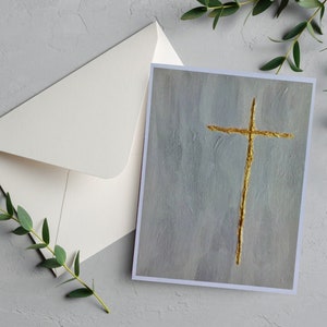 May include: A white card with a gold cross painted on a light blue background. The card is sitting on a gray surface with green leaves.