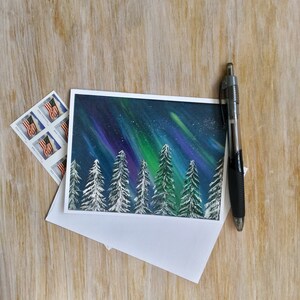 May include: A greeting card with a snowy forest scene under a vibrant green, blue, and purple aurora borealis. The card is blank inside for writing a message.