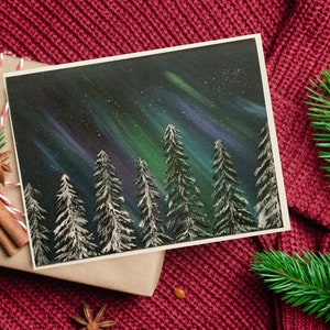 May include: A hand-painted card featuring a snowy forest scene under a vibrant aurora borealis. The sky is a deep blue with streaks of green and purple light. The trees are silhouetted against the sky, and their branches are covered in snow.