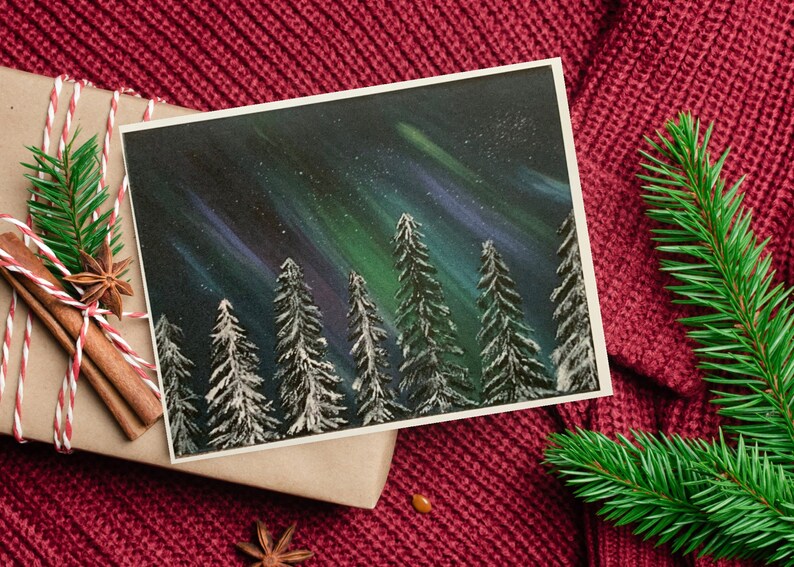 May include: A hand-painted card featuring a snowy forest scene under a vibrant aurora borealis. The sky is a deep blue with streaks of green and purple light. The trees are silhouetted against the sky, and their branches are covered in snow.