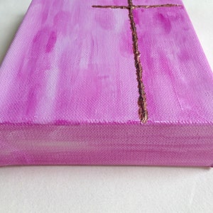 May include: A pink canvas painting with a gold cross in the center. The cross is made of a textured material that looks like glitter.