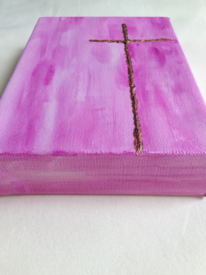 May include: A pink canvas painting with a gold cross in the center. The cross is made of a textured material that looks like glitter.
