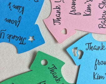 May include: A collection of colorful baby shower thank you tags in the shape of onesies. The tags are blue, pink, and green, with cut-out shapes like a star, heart, and footprints. Each tag has the text "Thank You from Kim's Baby Shower."