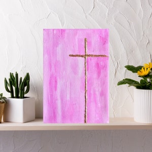 May include: A pink canvas painting with a gold cross in the center. The painting is on a shelf with two potted plants.