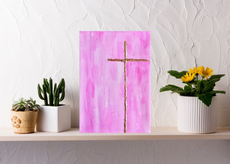 May include: A pink canvas painting with a gold cross in the center. The painting is on a shelf with two potted plants.