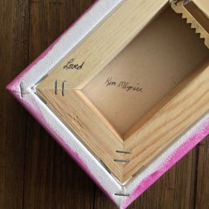 May include: The back of a pink and white canvas painting with a wooden frame. The painting is signed "Kim Mlyniec" and has the word "Loved" written on it.