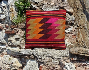 May include: A square throw pillow featuring a woven kilim pattern with horizontal stripes in red, orange, and black, accented by a central geometric zigzag design in shades of tan, orange, dark brown, and magenta against a deep red background.
