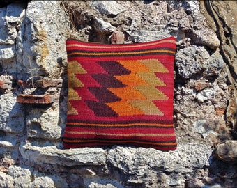 May include: A square throw pillow featuring a woven kilim pattern in shades of red, orange, dark brown, and gold. The design consists of horizontal stripes at the top and bottom with a central repeating geometric chevron motif across the surface.