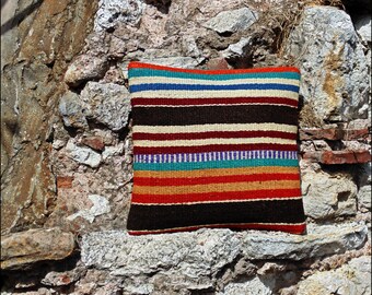 May include: A square throw pillow featuring a woven pattern of horizontal stripes in vibrant colors including teal, royal blue, cream, dark brown, red, and orange. The textile has a textured, coarse appearance with visible thread weave throughout.