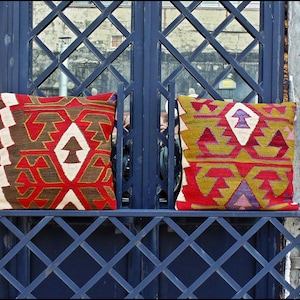 Puede incluir: Dos cojines cuadrados con complejos patrones geométricos en tonos rojo, marrón, amarillo y crema. Los cojines están colocados uno al lado del otro sobre un marco de celosía de metal azul oscuro, mostrando texturas tejidas y diseños.