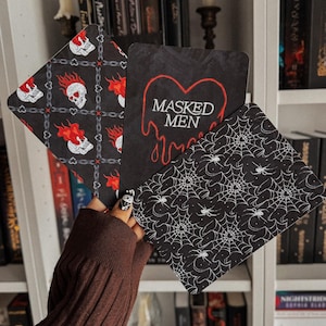 May include: Three Halloween-themed cards are held in a hand. One card features skulls with flames, another has the text "MASKED MEN", and the third displays a spiderweb design. The cards are black and white with red accents.
