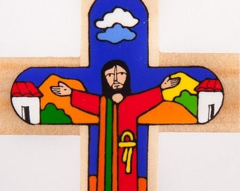 May include: A wooden cross with a colorful painted image of Jesus Christ with outstretched arms. The background features a blue sky with a white cloud, green trees, and two houses with red and yellow roofs.