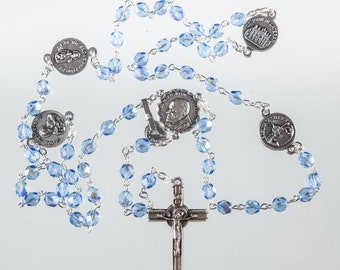 May include: A silver rosary with blue glass beads and a silver crucifix. The rosary features five silver medallions with religious imagery, including a depiction of Saint John Paul II.