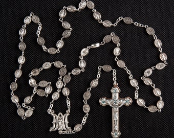 May include: A silver rosary with a silver crucifix featuring a depiction of Jesus Christ. The rosary beads are oval-shaped and have a raised design.
