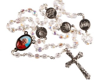 May include: A silver rosary with clear crystal beads and five silver medallions. One medallion features a portrait of Pope John Paul II, another features a depiction of the Holy Family, and another features a depiction of the Sacred Heart of Jesus. The rosary also includes a silver crucifix.