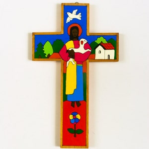 May include: A wooden cross with a colorful painted image of Jesus holding a lamb. The background includes a blue sky, green trees, a white dove, and a small white house with a red roof.