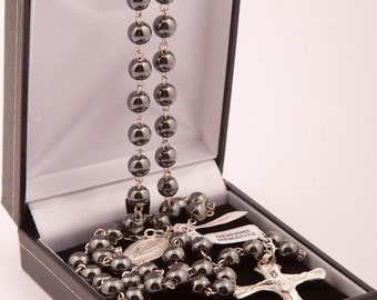 May include: A black hematite rosary with a silver cross and a silver medal in a black box. The box has a white interior and a label that reads "Genuine Hematite".