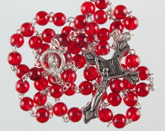 May include: A red rosary with a silver crucifix and a silver pendant featuring a depiction of Jesus. The rosary beads are made of glass and are strung on a silver chain.