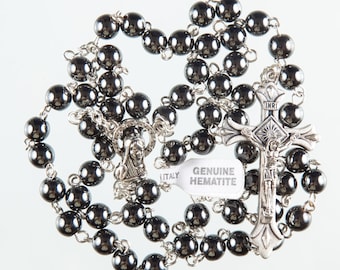 May include: A black hematite rosary with a silver crucifix and a silver charm featuring the Virgin Mary. The rosary beads are round and have a matte finish. The crucifix has a silver inscription "INRI" and a silver figure of Jesus Christ. The charm has a silver inscription "ITALY" and a silver figure of the Virgin Mary. The tag attached to the rosary reads "GENUINE HEMATITE".