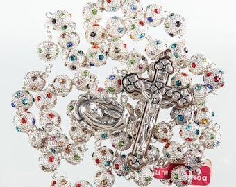 May include: A silver rosary with a silver crucifix and a silver pendant featuring the Virgin Mary. The beads are decorated with colorful rhinestones. The rosary is on a white background.