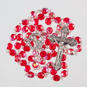 Può includere: Un rosario rosso con perline sfaccettate e un crocifisso d'argento. Il rosario presenta un elemento centrale d'argento con una raffigurazione della Vergine Maria. Le perline sono di un rosso vibrante.