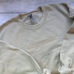 May include: A light beige sweatshirt with the text "binge reader" embroidered in a lighter beige color.