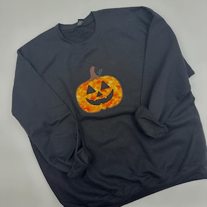 May include: Black crewneck sweatshirt featuring a jack-o'-lantern graphic. The pumpkin is orange and yellow with a smiling face and a brown stem. The sweatshirt is a solid black color, perfect for Halloween.
