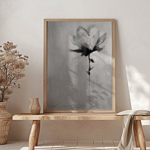 A monochromatic framed canvas print featuring a stylized flower in shades of gray. The flower is depicted with loose brushstrokes, creating a textured and painterly effect. The background is a soft, muted gray, providing a subtle contrast to the darker tones of the flower. The overall style is minimalist and modern, with a focus on the artistic expression of the floral subject. The print is displayed on a light-colored wooden bench, adding to the serene and calming atmosphere of the scene.
