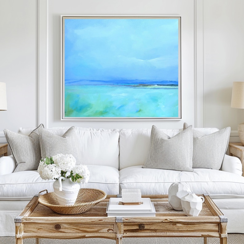 May include: A white couch with two gray pillows and a white vase with white flowers on a wooden coffee table. A large abstract painting of a blue and green seascape hangs on the wall above the couch.