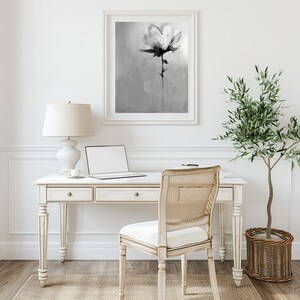A grayscale abstract floral painting of a single flower hangs above a white wooden desk.  A laptop, lamp, and chair are on the desk. A small olive tree in a woven basket sits on the floor next to the desk. The overall aesthetic is minimalist and modern.